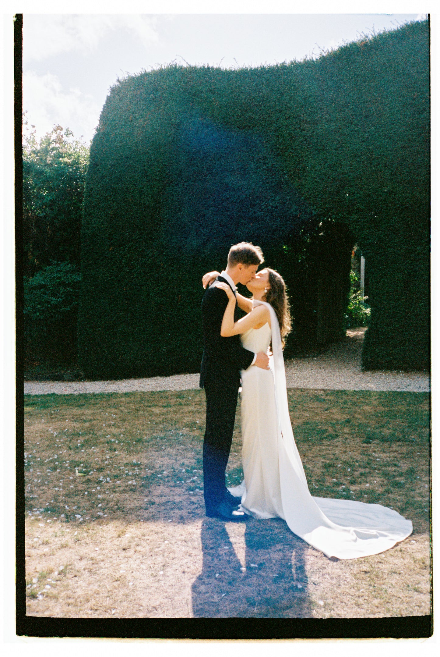 engaged couple embracing with trailing neck scarf in a garden setting with large hedges.
