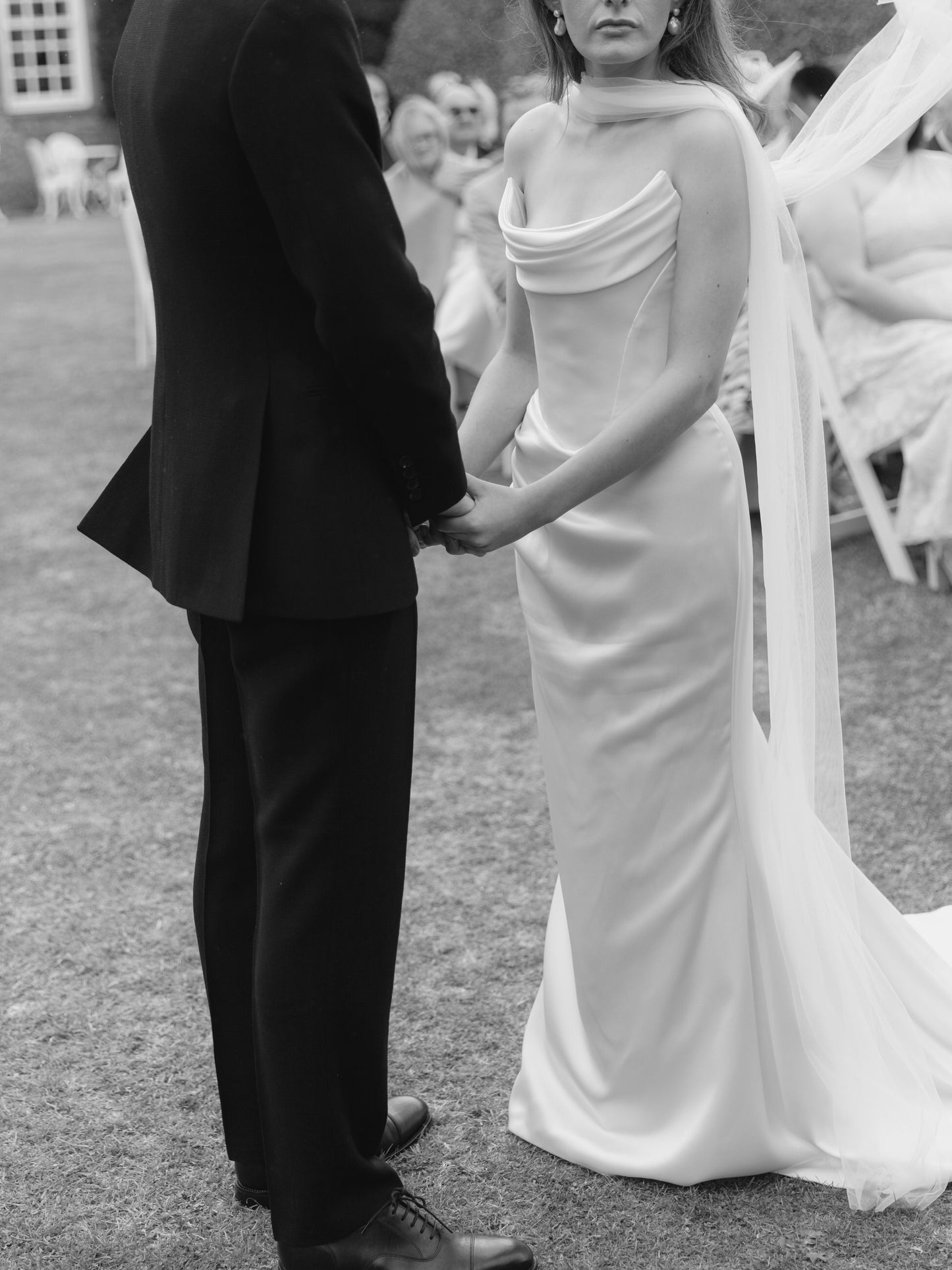 Black and white photo of an engaged holding hands outdoors with bride in cateye neckline Westwood gown and thin silk Tull bridal scarf