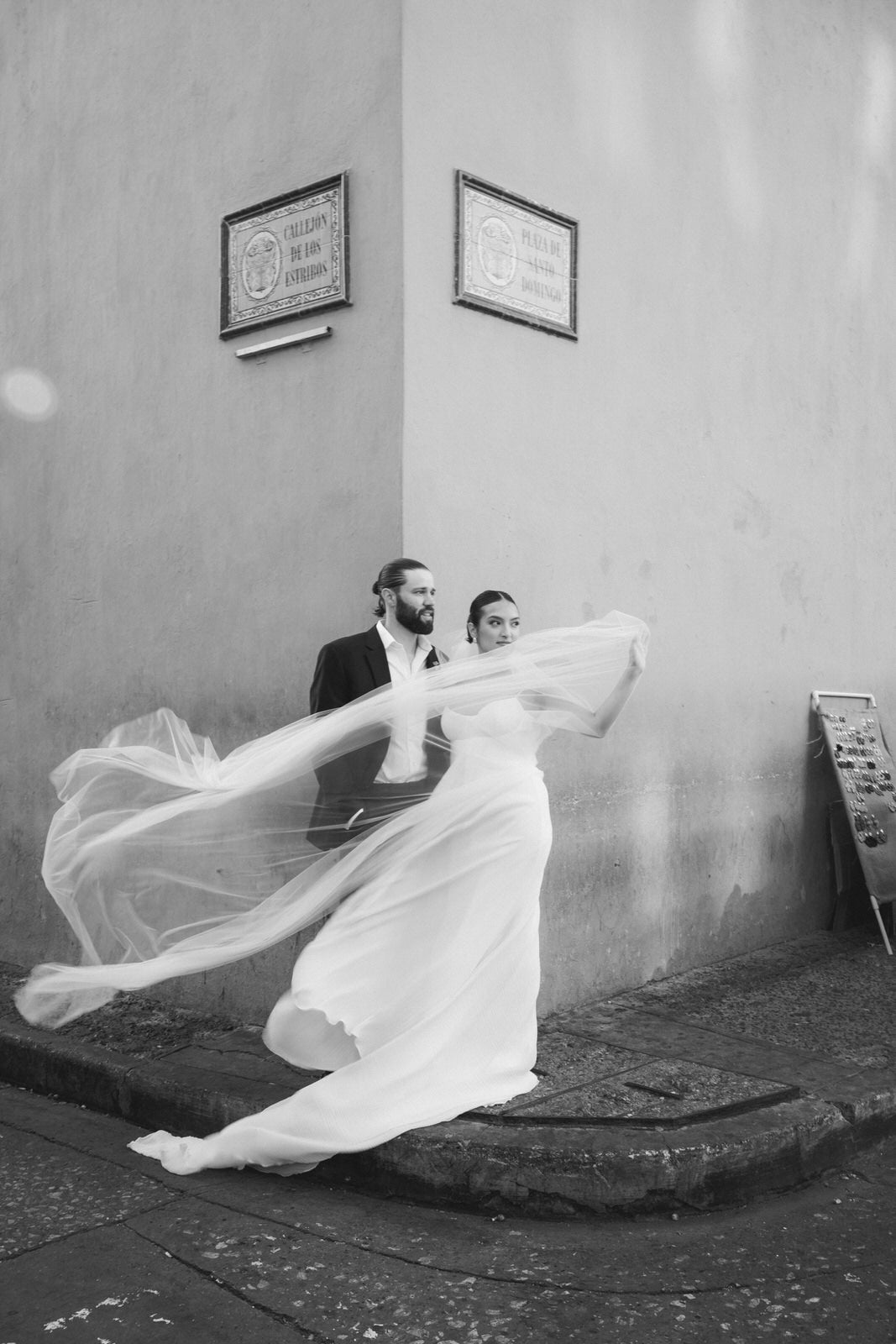 A black and white photograph of a bride and groom outdoors against a textured wall, with the bride's cathedral silk bridal veil dramatically spread