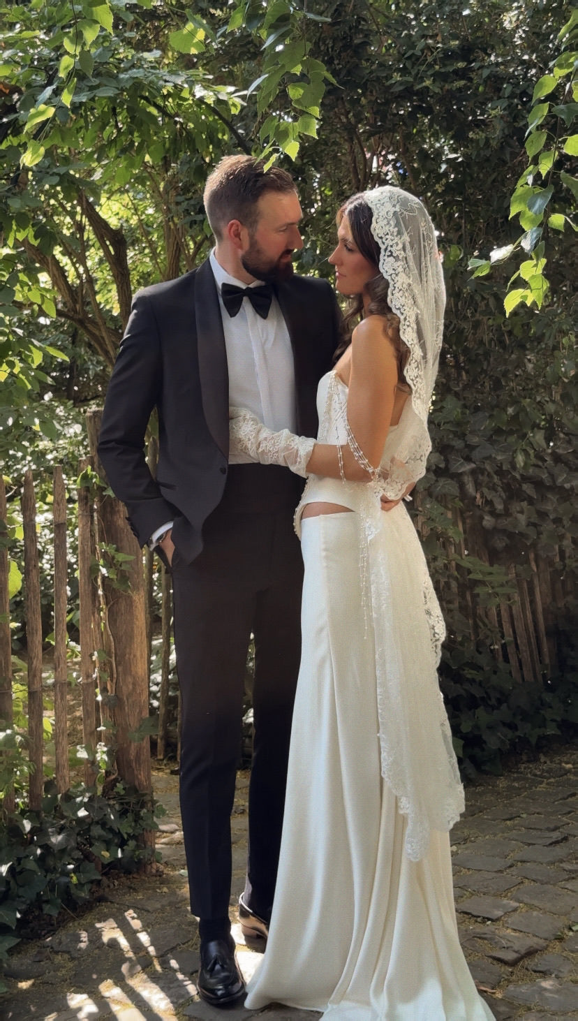 Bride in vintage inspired wedding attire and knee length mantilla veil standing outdoors with greenery in the background