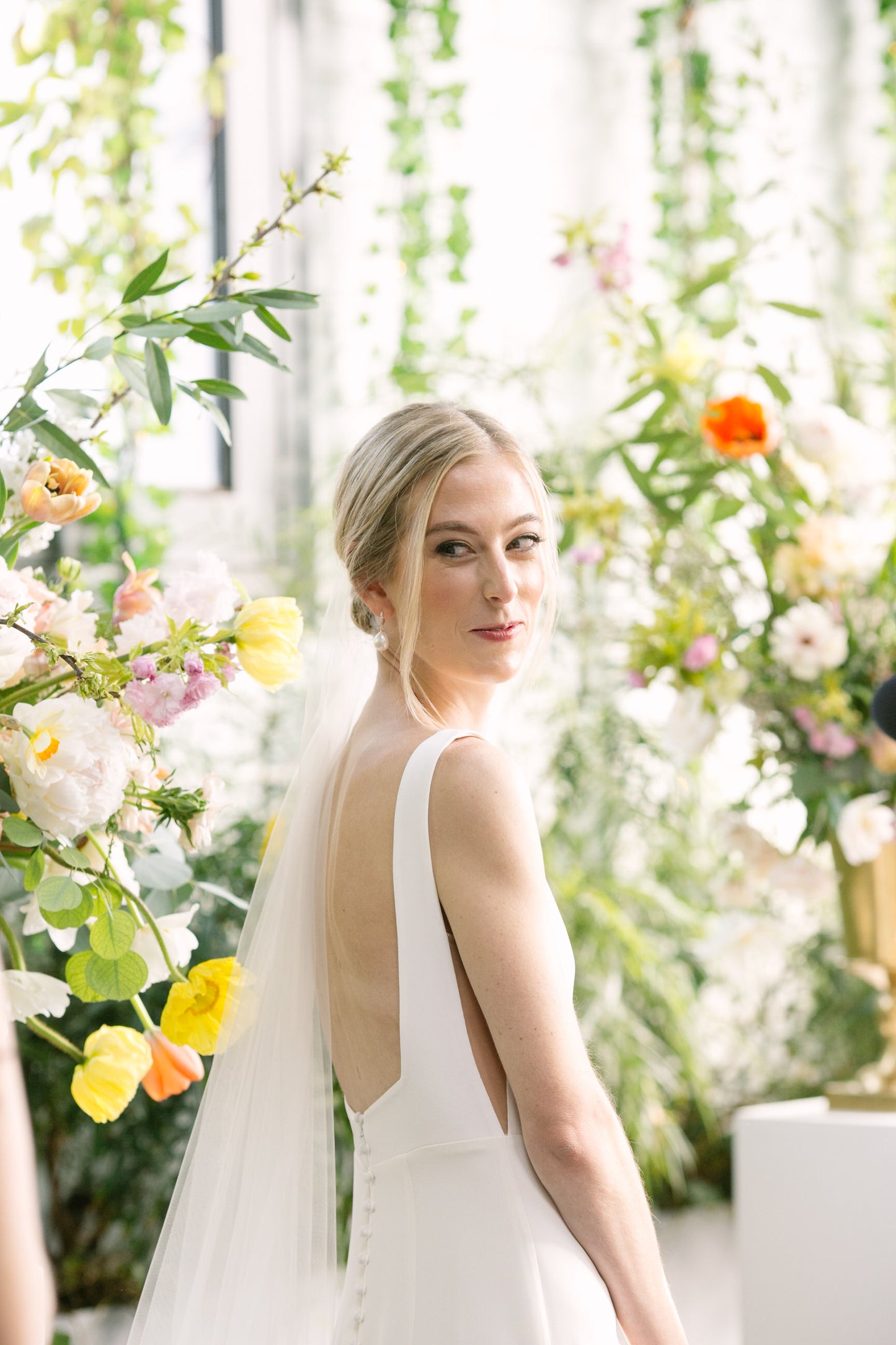 romantic off white cascade silk wedding veil over bun as bride looks over shoulder surrounded by flower arrangements