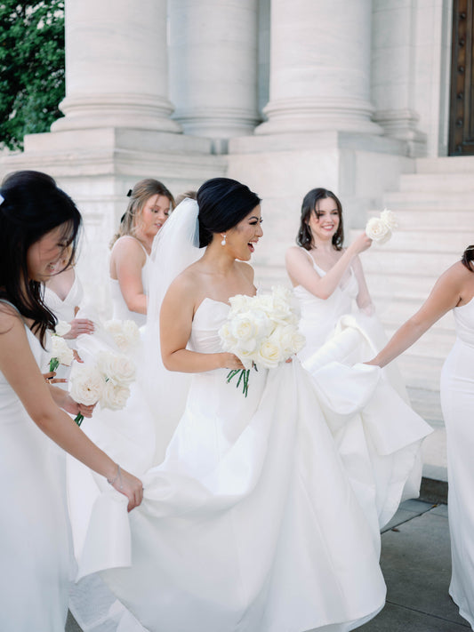 bridesmaids in white maxi dresses helping bride in ballgown and long wedding veil holding white bouquet