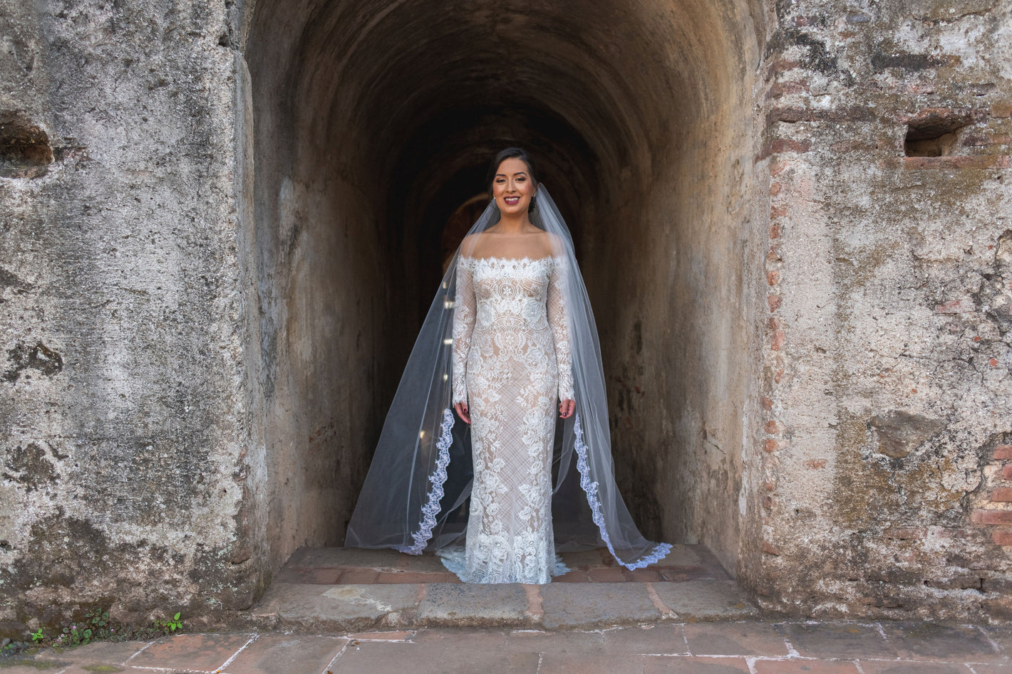 romantic cathedral length lace veil with lace trim and off the shoulder bridal dress as bride stands in Mexican archway