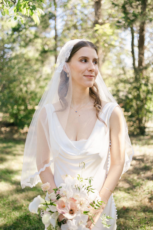 Woman in a white draped neck wedding dress with a Juliet cap wedding veil with thin scallop trim holding a bouquet of flowers outdoors.