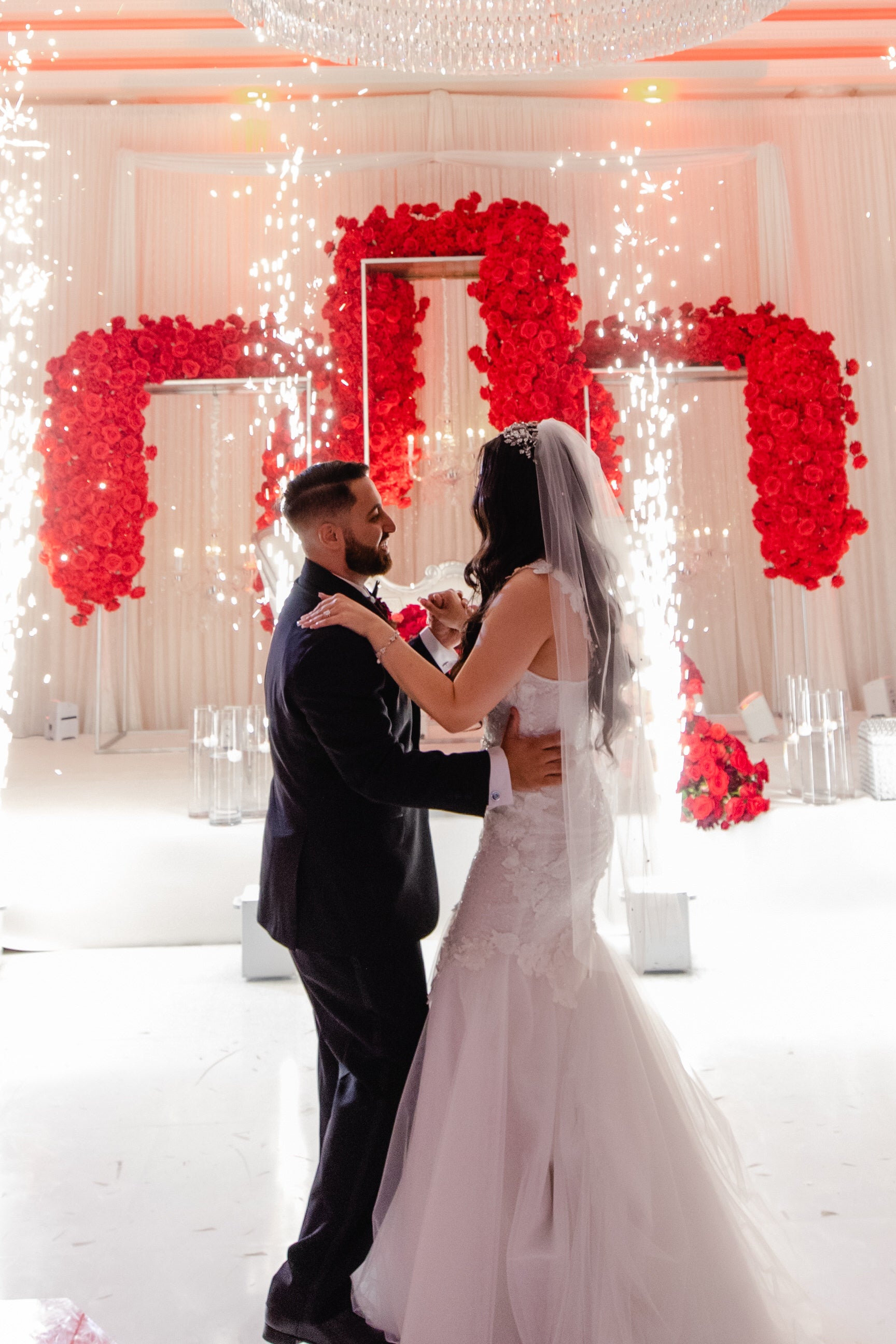 Persian couple with bride wearing fingertip length rhinestone wedding veil in downdo hairstyle with red rose arches in background
