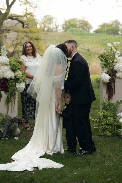 A bride in a white vivienne Westwood old glam wedding gown and puffy knee length veil kissing her groom in a black suit outdoors, with a young ring bearer standing beside them 
