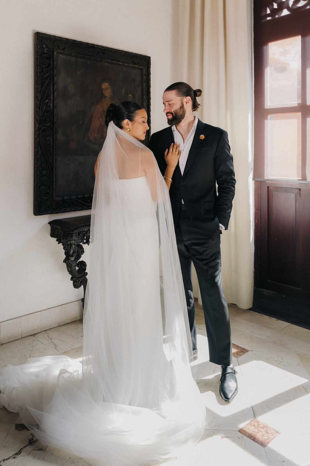 A bride in a white spaghetti strap wedding gown with a long raw edge cathedral veil and a groom in a black suit standing indoors beside a window and framed artwork.