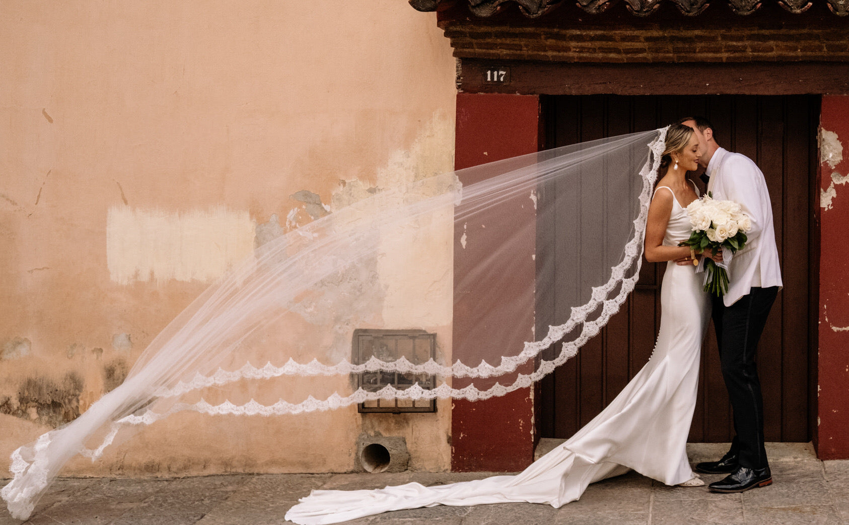 Spanish style mantilla eyelash lace royal length wedding veil on bride and groom in Spanish doorway with veil blowing