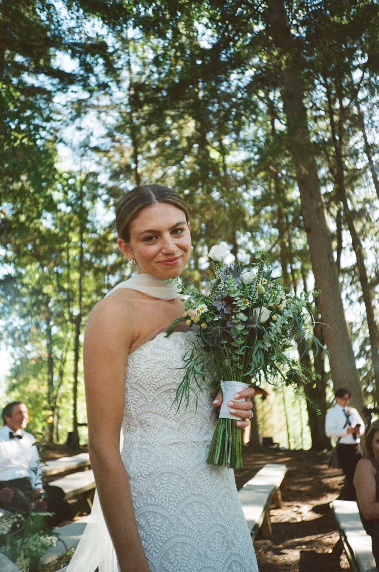 Woman in a lace wedding dress and narrow bridal scarf holding a bouquet of wildflowers outdoors with trees in the background