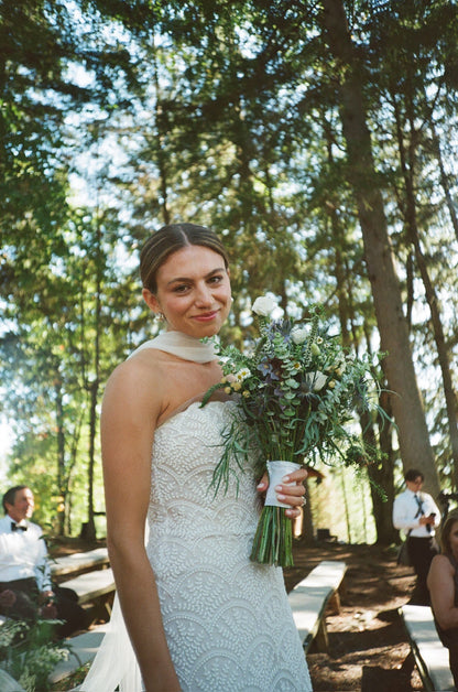 Woman in a lace wedding dress and narrow bridal scarf holding a bouquet of wildflowers outdoors with trees in the background
