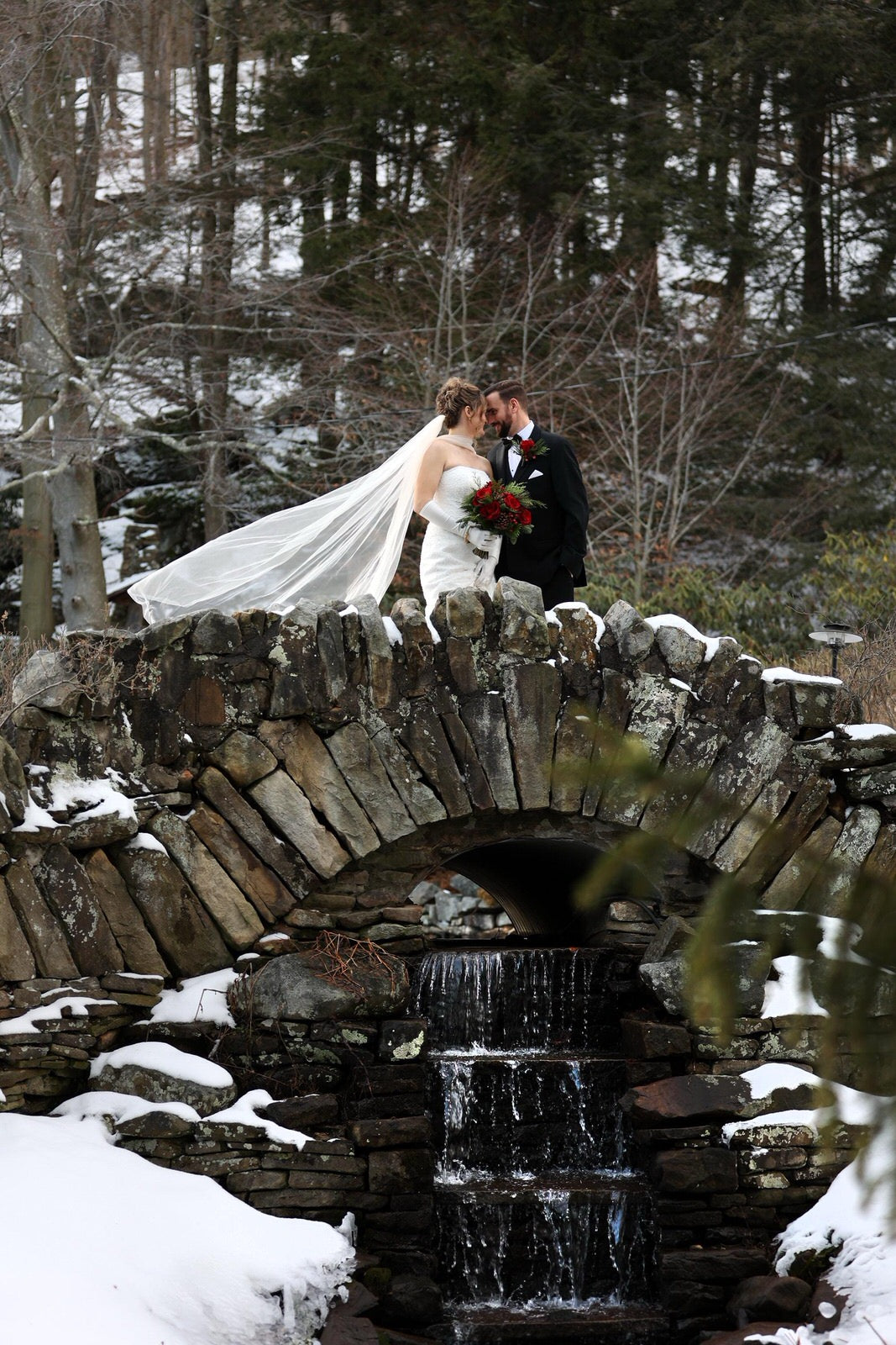 Bride in scarf and veil standing on a stone bridge over a small waterfall in a snowy forest.