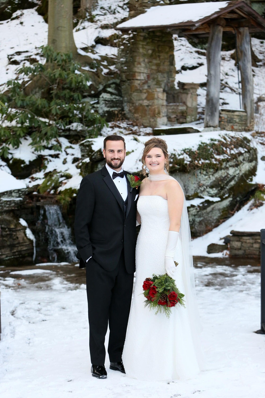 Bride in scarf from tulle and matching gloves in satin gloves standing in a snowy landscape with a gazebo and waterfall in the background.