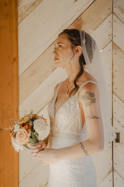 A jewish bride with tattoos in a white lace wedding dress and sheer veil holds a peach and white floral bouquet, standing in front of a rustic wood-paneled wall.