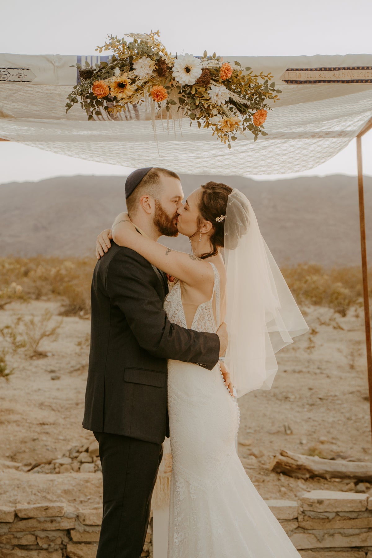 A bride in a white lace wedding gown and veil and a groom in a black suit kiss under a floral chuppah outdoors in a desert.