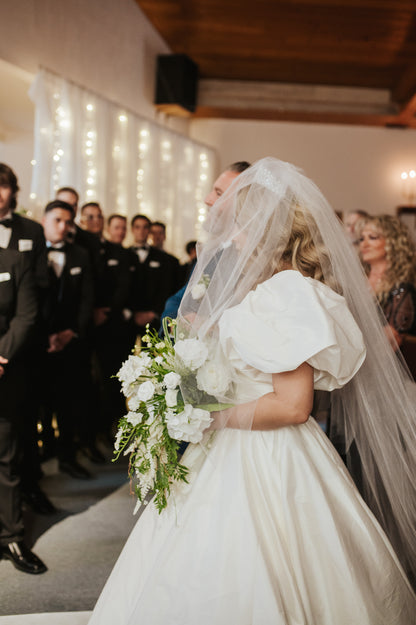 Bride in a white  puff sleeve wedding dress with a long cathedral veil holding a bouquet, surrounded by guests in formal attire.