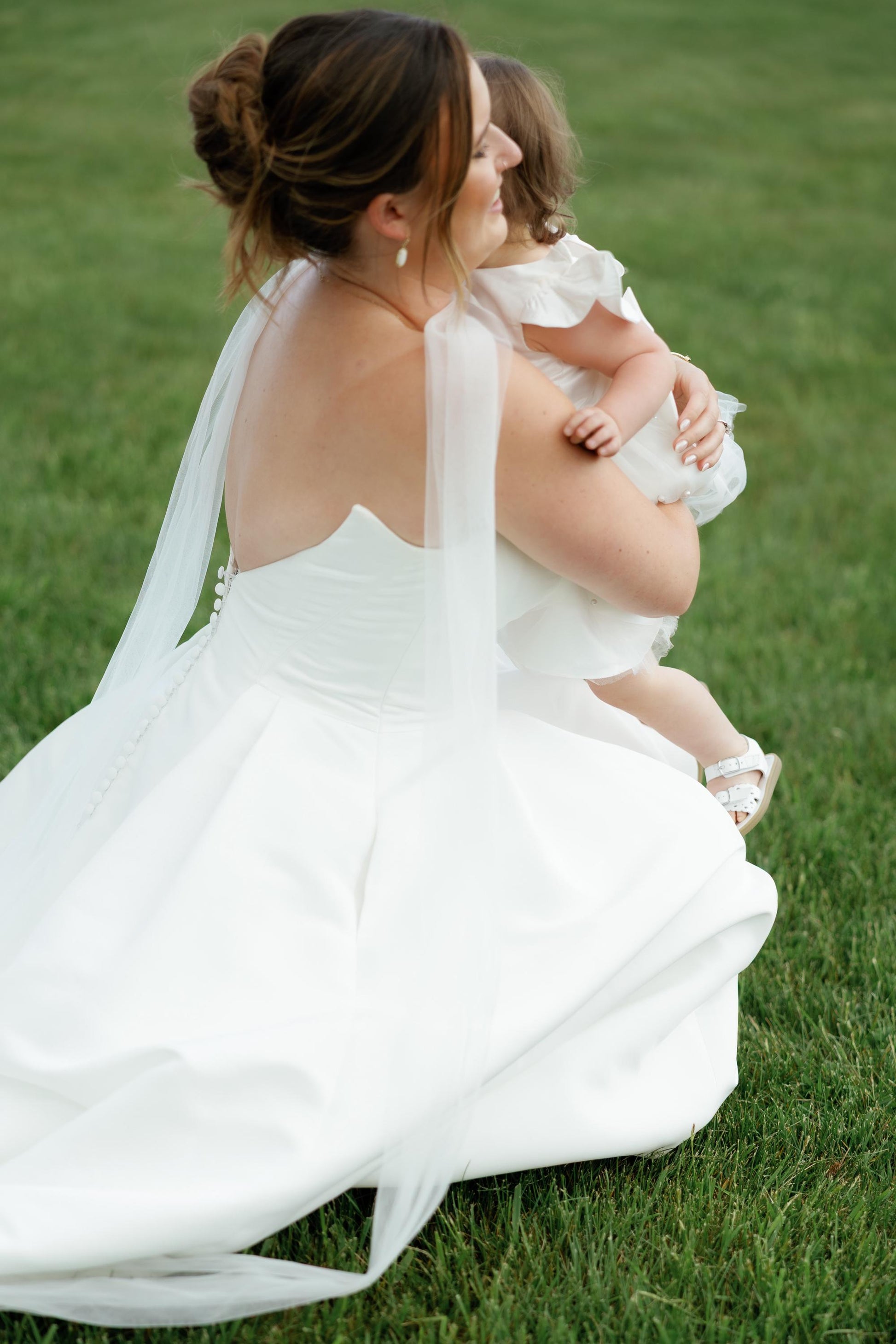 bride in a white strapless wedding dress wearing DIY bridal tulle scarf and holding a flower girl on grass