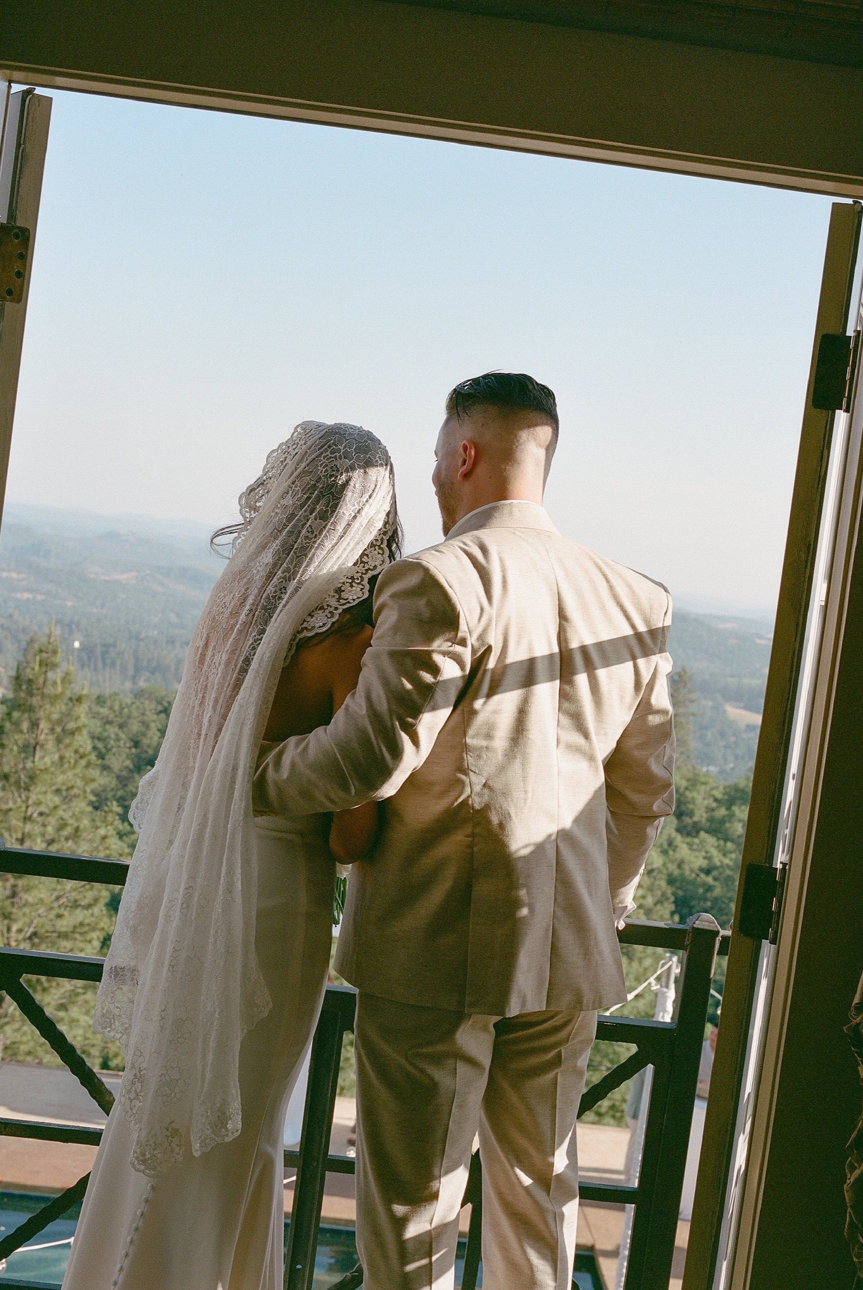 bride and groom look out balcony with bride in mid length lace mantilla veil on top of head and sheath gown