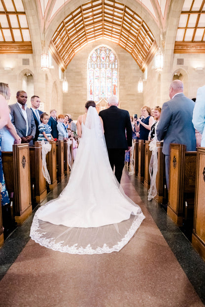 custom long cathedral length wedding dress in ivory with lace trim on bride as she walks down aisle of church with Dad