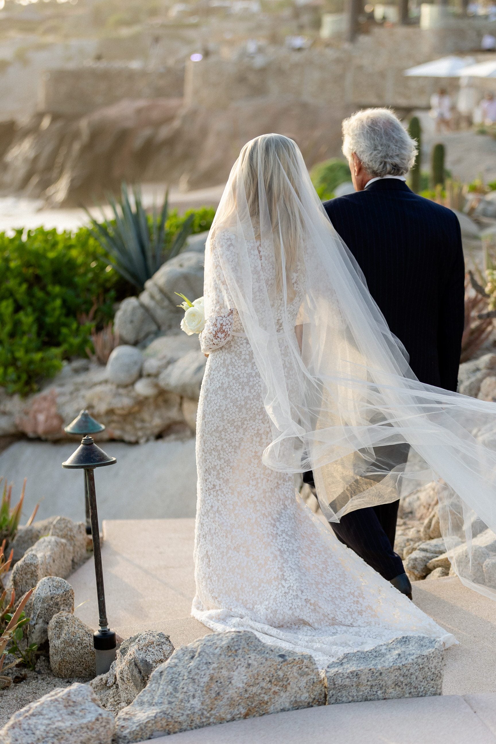 bride walking down path with dad in long sleeved lace bridal dress and blowing cathedral length wedding veil