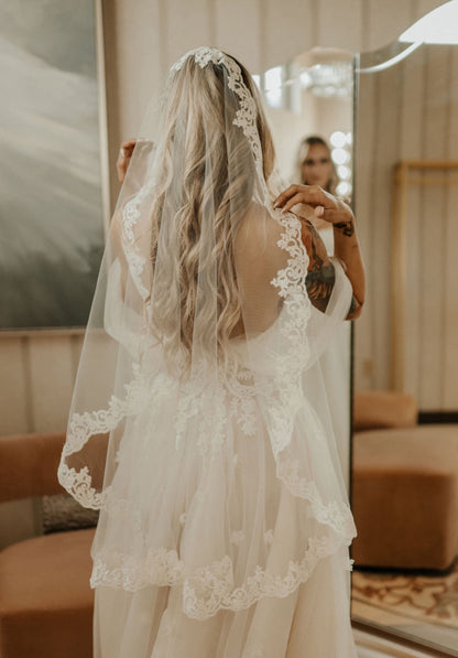 romantic circle mantilla cut wedding veil with ivory French lace trim on bride in soft tulle wedding gown looking in mirror