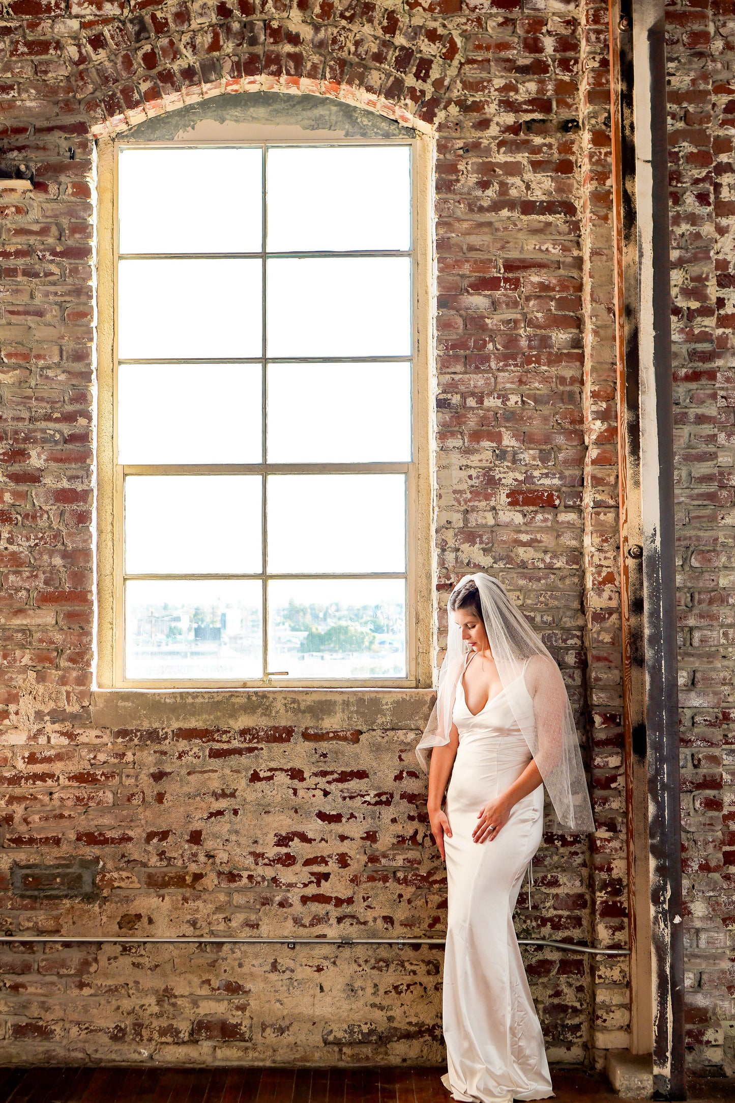 bride standing by window at red brick modern studio wearing a mid length ivory polka dot bridal veil